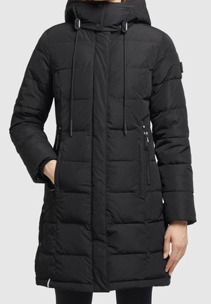 Long manteau d'hiver matelassé noir avec capuche, cordons, poches latérales zippées et boutons-pression sur les côtés, porté par une personne avec une main dans la poche.