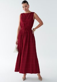 Rød maxikjole med en struktureret overdel, smocket talje og tekstureret krøllede stof. Har et ærmeløst design og elegant, flydende nederdel.