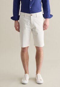 Weiße Baumwollshorts mit geradem Schnitt, mit Knopfverschluss und Taschen. Kombiniert mit einem blauen Langarmhemd und weißen Turnschuhen.