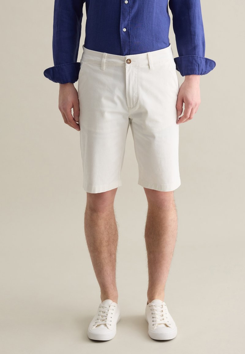 Weiße Baumwollshorts mit geradem Schnitt, mit Knopfverschluss und Taschen. Kombiniert mit einem blauen Langarmhemd und weißen Turnschuhen.