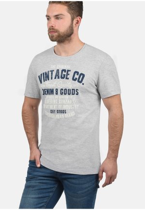 Siva katirana majica s kratkim rukavima koja sadrži tamnoplavi ispisani tekst "Vintage Co." i druge detalje brendiranja na prednjem dijelu.