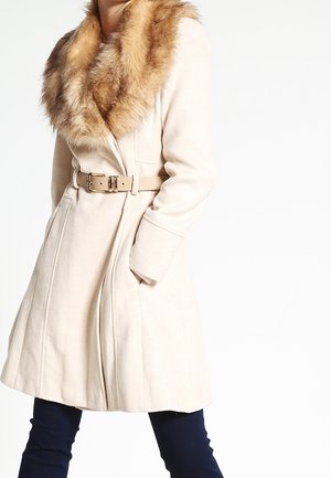 Femme portant un manteau beige ceinturé avec un grand col en fausse fourrure marron et un pantalon foncé sur un fond blanc.