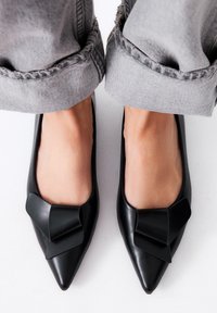 Bailarinas de cuero negro con punta afilada que presentan un gran lazo en la parte delantera, combinadas con pantalones grises con puños. Textura suave y diseño elegante.