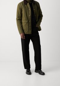 Veste utilitaire vert olive avec un col en velours côtelé marron, des boutons-pression et deux poches avant, associée à un pantalon noir à jambes larges et des chaussures noires.