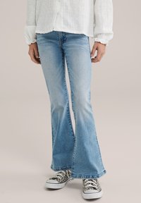 Zúžené modré džínsy s rozšírením na spodku a svetlou prateľnou úpravou, so tesným strihom v oblasti bokov a širším lemom. Skombinované s čierno-bielymi teniskami so vzorom.