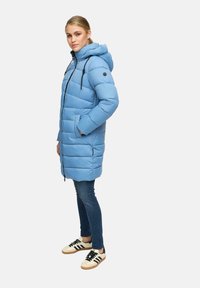 Vrouw in een lichtblauwe gewatteerde winterjas met capuchon, donkere spijkerbroek en witte sneakers met zwarte strepen, staande in zijaanzicht.