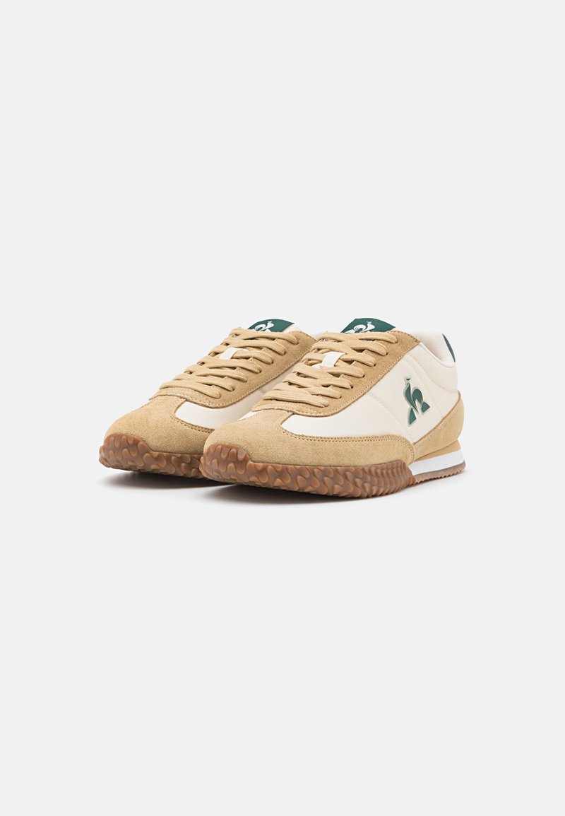 Baskets en daim beige et en cuir crème avec des accents verts, semelle en caoutchouc texturée et lacets plats. Présente un logo sur le côté et un col rembourré.
