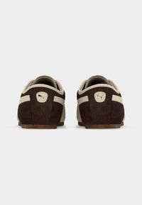 Ζευγάρι καφέ και μπεζ αθλητικά παπούτσια Puma, φωτογραφημένα από πίσω σε απλό λευκό φόντο, με έμφαση στο λογότυπο στη φτέρνα.