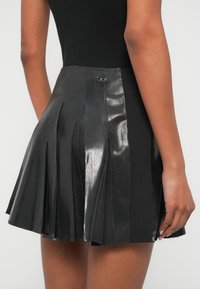 Juoda klostuota sijonas iš blizgios dirbtinės odos medžiagos, su megzta juosmens juosta ir metaline logotipo detale gale.