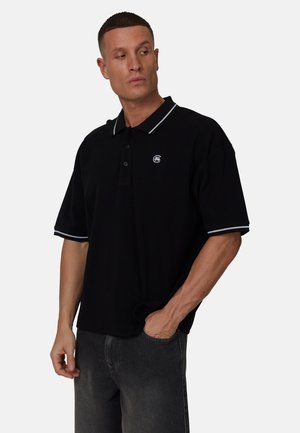 Polo noir à manches courtes, avec des rayures blanches sur le col et un petit logo sur la poitrine. Le tissu semble texturé et la coupe est décontractée.