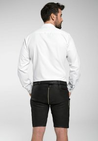 Weißes Langarmhemd mit Knopfmanschetten, getragen mit schwarzen Shorts mit gelben Nähten und Seitentasche, von hinten gezeigt.