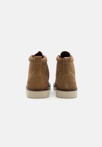 Botas de tobillo de ante marrón con puntera redonda, costuras mínimas y suela de goma beige gruesa. Asas en la parte trasera para facilitar el calzado.