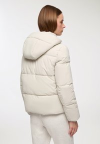 Kobieta w jasnobeżowej pikowanej kurtce z kapturem, pokazana od tyłu, na jednolitym białym tle.
