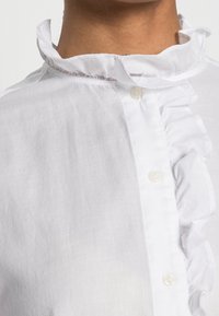 Blusa bianca con colletto arricciato, apertura con bottoni e texture liscia. Il tessuto appare leggero e leggermente trasparente con un sottile luccichio.