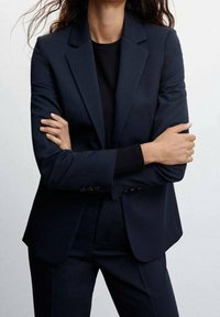 Blazer bleu marine ajusté avec une coupe slim, des revers crantés et une texture lisse. Porté avec un pull noir ajusté en dessous.