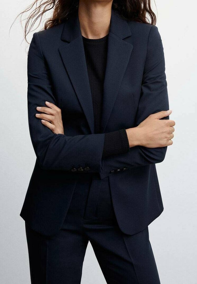 Blazer bleu marine ajusté avec une coupe slim, des revers crantés et une texture lisse. Porté avec un pull noir ajusté en dessous.