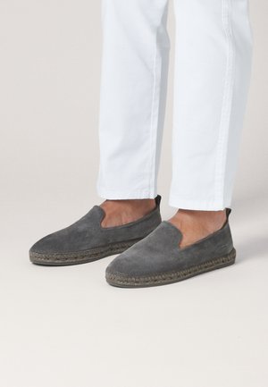 Fødder iført grå ruskind slip-on espadriller kombineret med hvide bukser mod en neutral baggrund.