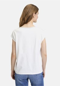 Weißes kurzärmeliges Oberteil mit Rundhalsausschnitt und grauen Nähten, kombiniert mit blauen Jeans. Aus weichem Baumwollmaterial, lockere Passform, glatte Textur.