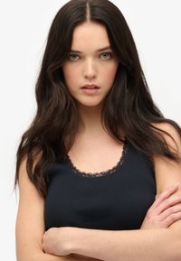 Sort ribbet tanktop med blonder trim langs halsudskæringen og ærmegabene. Stofserien ser glat og tætsiddende ud. Modellen står med armene krydset.