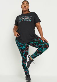 Musta t-paita, jossa teksti "OLE ITSESI AKTIIVINEN"; särmikkäät leggingsit mustassa, tealissa ja burgundissa; täydentää mustat urheilukengät.