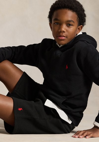 Chlapec oblečený v čiernej mikine s kapucňou a šortkách s červenými logami, sediaci s jednou rukou opretou o podlahu na jednoduchom svetlom pozadí.