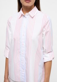 Button-up shirt with a white base featuring vertical stripes in red, blue, and yellow. Collared with rolled sleeves and button closure.