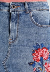 Jupe en denim avec un délavage bleu, featuring des broderies florales complexes en rouge, rose et bleu, mettant en valeur des détails texturés sur le tissu.