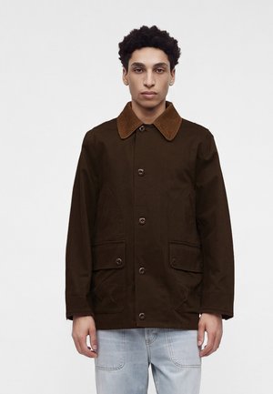 Junger Mann mit lockigem Haar, der eine dunkelbraune, geknöpfte Jacke mit kontrastierendem Kordkragen und hellblaue Jeans vor einem weißen Hintergrund trägt.