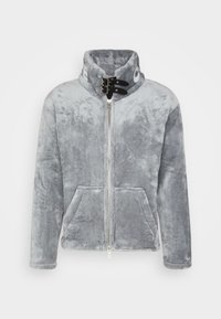 Szara kurtka polarowa z wysokim kołnierzem, srebrnym zamkiem błyskawicznym, dwoma przednimi kieszeniami i czarnymi akcentami w postaci pasków. Miękka tekstura z puszystym wykończeniem.