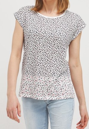 Femme portant un chemisier blanc à manches courtes avec de petits motifs floraux noirs et rouges, associé à un jean bleu clair.