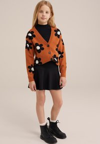 Cárdigan de punto naranja con un patrón floral en blanco y negro, combinado con una falda negra y botas de combate negras, que incluyen calcetas a rayas blancas.