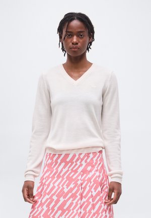 Γυναίκα με κοντά dreadlocks, ντυμένη με λευκό πουλόβερ με V λαιμόκοψη και φούστα με ροζ και λευκό μοτίβο, στέκεται μπροστά σε ένα απλό φόντο.