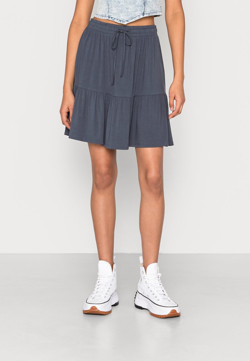 Femme portant une jupe longue grise foncée à volants jusqu'aux genoux avec une taille à cordon, des baskets montantes blanches et un haut en denim clair court.