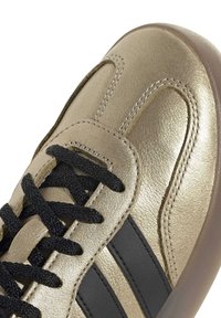Acercamiento a una zapatilla de oro con cordones negros y rayas negras en el costado, con costuras detalladas y una suela marrón.