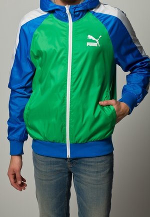 PUMA větrovky s zeleným předním dílem a modrými rukávy, vybavené bílým zipem a logem, vyrobené z hladkého, lehkého materiálu.