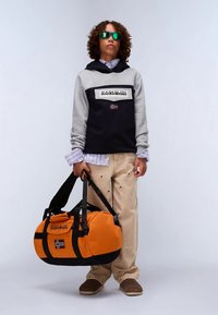 Tini, göndör hajjal és napszemüvegben, narancssárga sporttáskát tart, szürke és fekete kapucnis pulóvert, csíkos pólót, bézs nadrágot és barna cipőt visel.