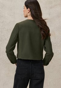 Dark green cropped jacket in soft fabric, featuring long sleeves and a clean neckline. Paired with dark denim jeans with subtle stitching.