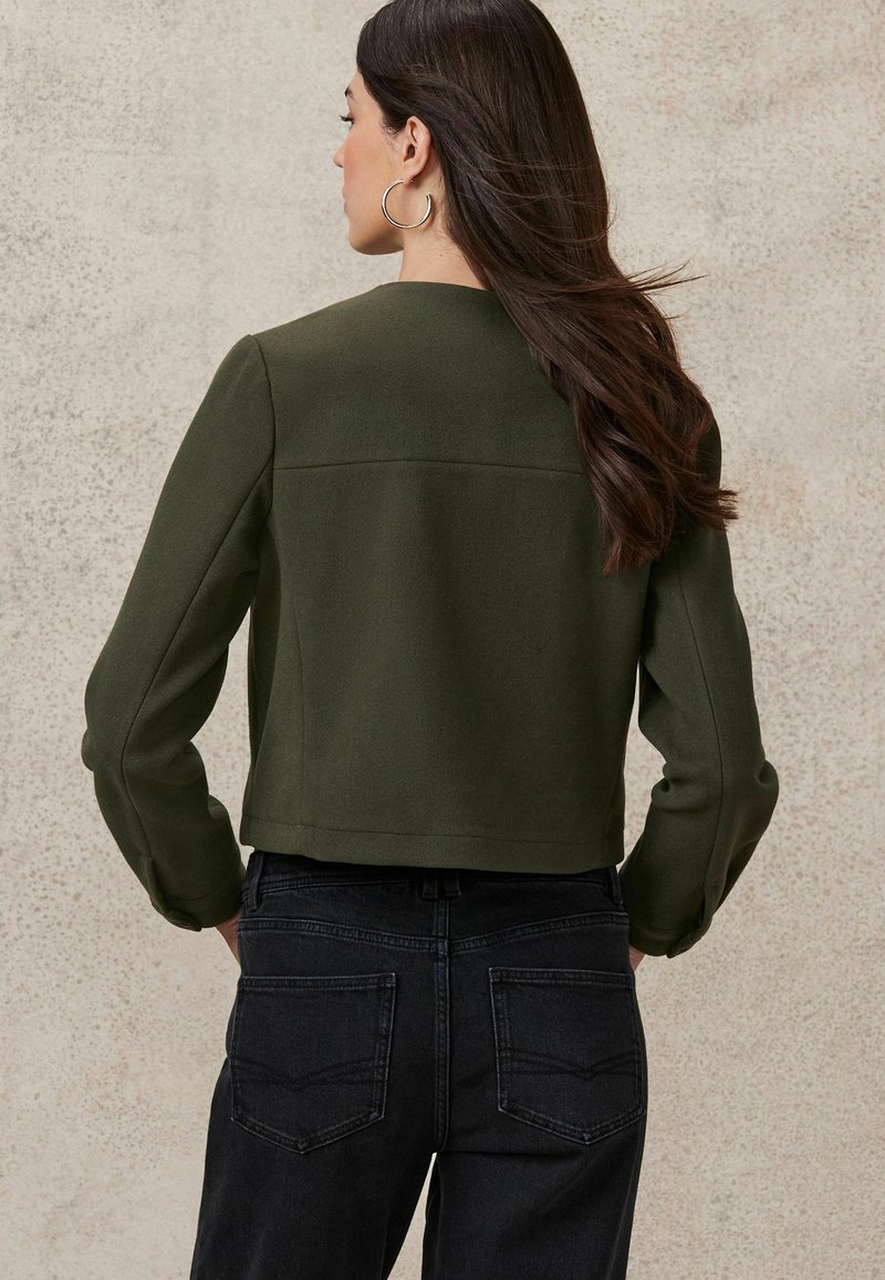 Dark green cropped jacket in soft fabric, featuring long sleeves and a clean neckline. Paired with dark denim jeans with subtle stitching.