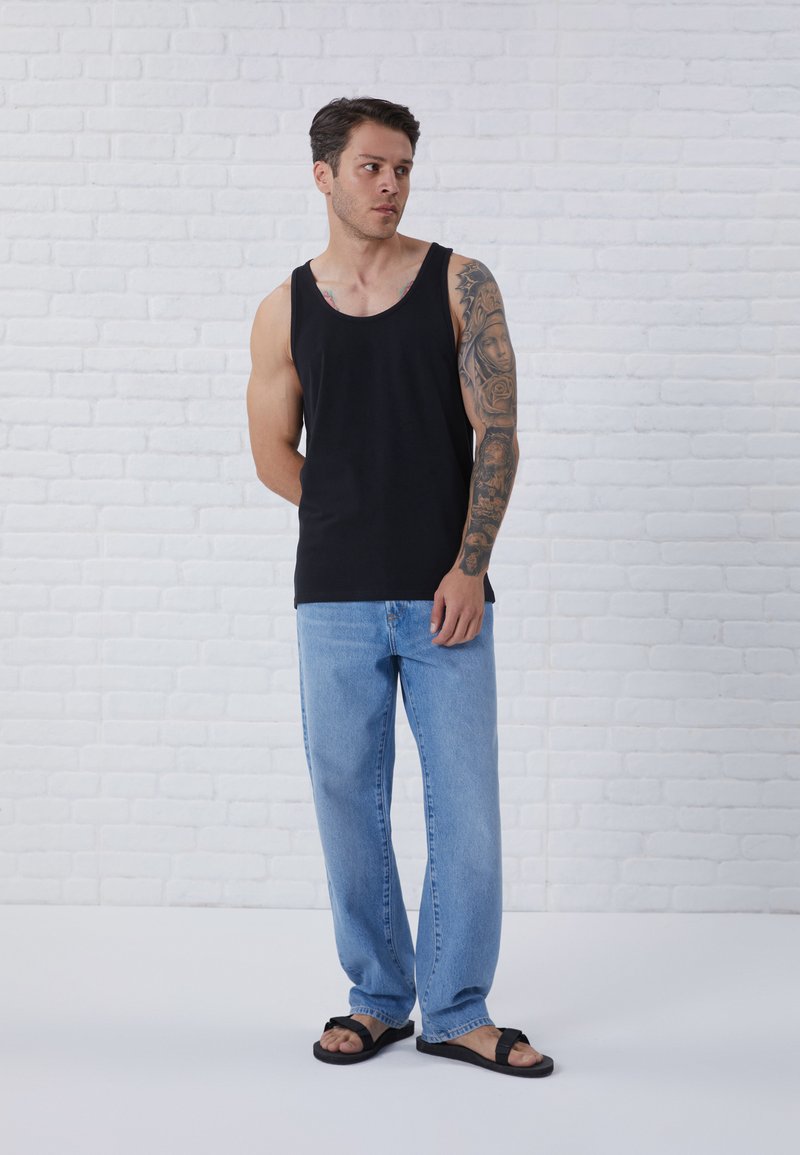 Zwarte mouwloze tanktop, lichtblauwe recht gesneden spijkerbroek en zwarte slippers. Model heeft een tatoeage op de arm en staat tegen een witte muur.