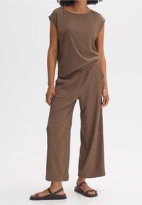 Kvinna i brunt texturerat ärmlöst linne och vida byxor med mörkbruna sandaler, står mot en enkel bakgrund.
