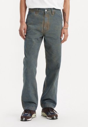 Personne portant un jean bleu délavé à jambes droites, une chemise blanche et des chaussures de course marron, bleu et crème, debout devant un fond uni.