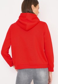 Felpa rossa con cappuccio e lacci tono su tono, vestibilità comoda e tasca a marsupio. Texture liscia, polsini e orlo a costine, abbinata a jeans grigi sbiaditi.