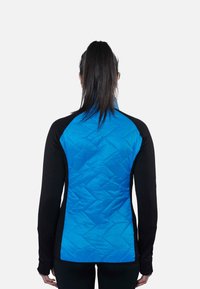 Femme aux longs cheveux bruns attachés, portant une veste noire avec un panneau arrière matelassé bleu vif, debout de dos sur un fond uni.