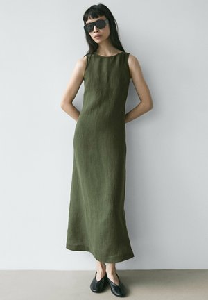 Donna che indossa un vestito midi verde oliva senza maniche, scarpe nere piatte e grandi occhiali da sole neri, in piedi contro una parete grigia chiara liscia.