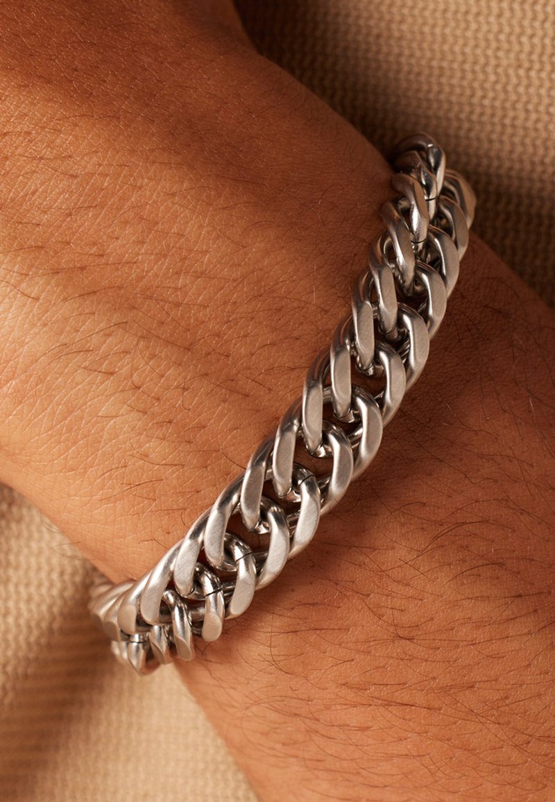 Zilveren metalen kettingarmband met in elkaar grijpende schakels, gepolijste afwerking en een stevige constructie, gedragen om een pols voor een textuurachtige achtergrond.