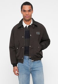 Juodas bomber jacket su užtrauktuku priekyje, spaudėmis ir šoninėmis kišenėmis. Yra raištis apykaklėje ir rankogaliuose, kairėje krūtinėje yra logotipo pleistras.