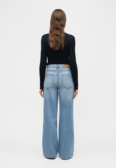 Άτομο με μακριά, κυματιστά καστανά μαλλιά, ντυμένο με μαύρη μπλούζα με μακριά μανίκια και φαρδιά τζιν παντελόνια ανοιχτού μπλε χρώματος, κοιτάζοντας προς τα πίσω σε απλό φόντο.