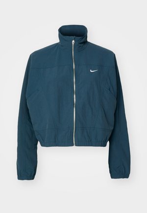 Giacca cropped blu navy con colletto alto, chiusura con zip, polsini elasticizzati e logo Nike bianco sul petto sinistro. Texture morbida.