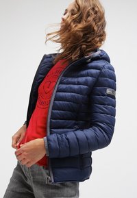 Granatowa kurtka puchowa z poziomym pikowaniem, kapturem i zapięciem na zamek, zestawiona z czerwoną koszulką z grafiką i szarymi dżinsami.