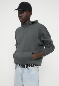 Ciemnoszara bluza z kapturem, posiadająca kieszeń z przodu i luźny krój, zestawiona z jasnoniebieskimi dżinsami oraz czarnym paskiem z ćwiekami. Czarna czapka dopełnia stylizację.